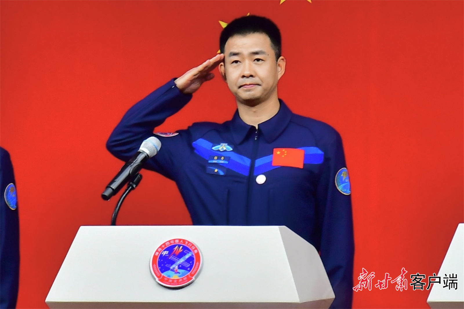 在见面会现场,三名航天员身着深蓝色航天服,背靠五星红旗,向全国人民